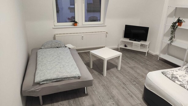 A modern, minimalist bedroom with a bed, a small white table, a TV on a stand, and a radiator. - Beautiful apartment on the roof floor for 5 people (Zeitz)