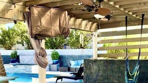 Área externa de piscina com espaço coberto para sentar, espreguiçadeiras e uma mesa de madeira.