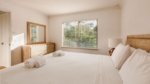 Ein Hotelzimmer mit einem großen Bett, einem hölzernen Kopfteil, einem Nachttisch mit Lampe, einem Spiegel und einem Fenster mit Jalousien.
