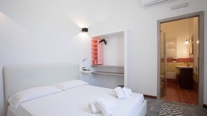 A hotel room with a bed, white linens, a pink shelf with a towel, a bathroom visible through an open door, and a patterned floor. - Downtown poolfront room with private veranda (Oristano)