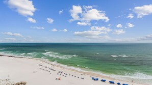 A beach with a clear sky, calm sea, and a sandy shore with blue umbrellas and lounge chairs. - Phoenix Orange Beach!  Lazy River and Slide !  New Rental!! (Orange Beach)