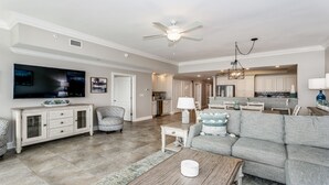 A spacious living room with a large sofa, a television, and a dining area with a kitchen in the background. - Phoenix Orange Beach!  Lazy River and Slide !  New Rental!! (Orange Beach)