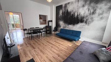 Ein modernes Wohnzimmer mit einem blauen Sofa, Holzboden, einem Essbereich mit Stühlen und einem großen Wandbild mit Waldmotiv.