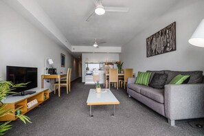 A modern living room with a grey sofa, a coffee table, a TV, and a dining area with chairs and a table. - ZEN CITY & SEA - Executive 1BR Suite (Darwin)
