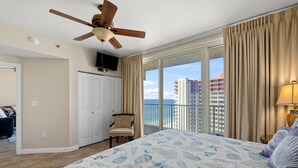 Ein Hotelzimmer mit einem großen Bett, einem Deckenventilator, einem Sessel, einem Fernseher und einem Balkon mit Blick auf das Meer und Gebäude.