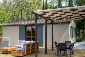 Un espace extérieur aménagé, abrité et doté d’un banc et d’une table en bois, d’une pergola illuminée et d’un bâtiment dont le toit est recouvert de tuiles.