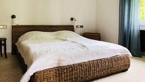 Una cama bien hecha con cabecera de mimbre, una lámpara de noche y una mesita pequeña.