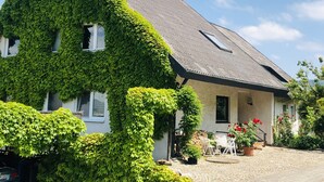 Una casa con techo empinado cubierto de hiedra, un patio de piedra y un pequeño espacio exterior con asientos.