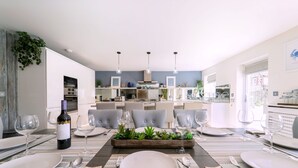 A modern dining area with a central table set for four, surrounded by chairs. There are wine glasses and a bottle of wine on the table. The room has a neutral color palette with a mix of natural and artificial lighting. - Drifters, Thornham, Norfolk (Thornham)