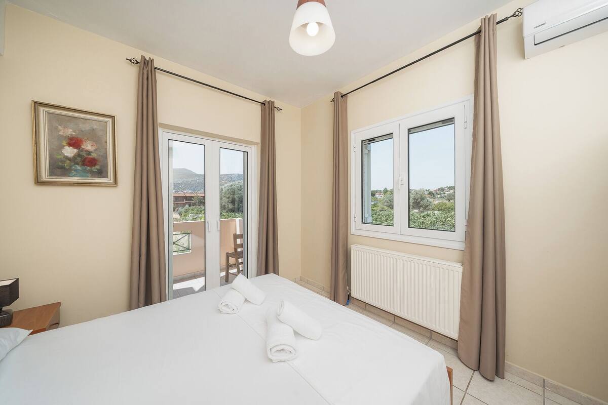 Une chambre d’hôtel avec un lit, un radiateur, une fenêtre avec des rideaux, une porte donnant sur un balcon et un tableau au mur.