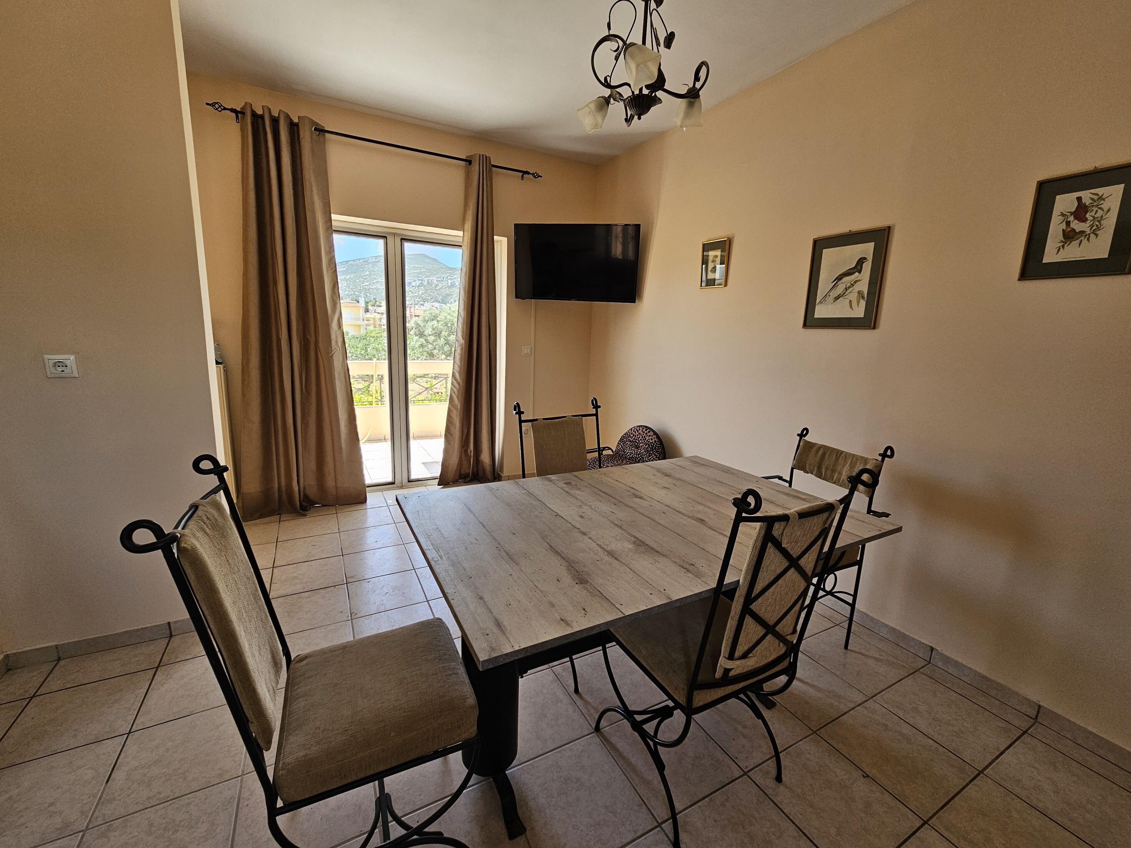 A dining area with a wooden table and chairs, a TV mounted on the wall, and a view of the outdoors through a sliding door.