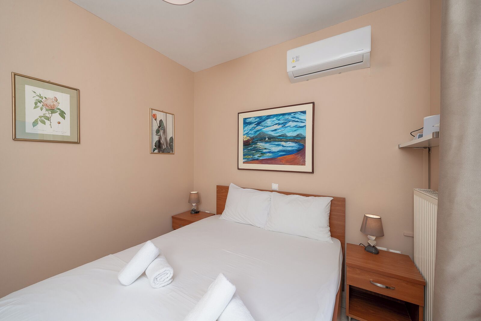 Quarto de hotel com uma cama, duas mesas de cabeceira, ar-condicionado e obras de arte emolduradas nas paredes.