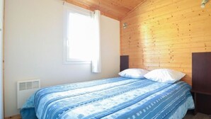 A bedroom with a wooden ceiling, a window with white curtains, a bed with blue bedding, and a wall-mounted heater.