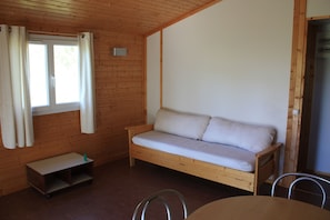 A wooden cabin room with a bed, a small table, and a window with curtains.