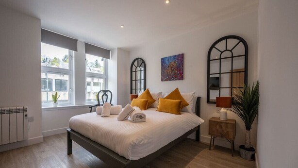 A modern bedroom with a large bed, two windows, a radiator, a bedside table, and a potted plant. - Mrs Watermelon Apartment (Fort William)