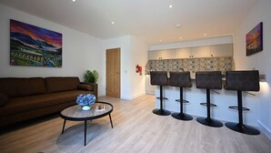 A modern living room with a brown sofa, a coffee table with a vase, and a kitchen area with a bar stool seating. - Mrs Watermelon Apartment (Fort William)