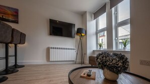 A modern living room with a wooden floor, a round coffee table with a remote control, a potted plant, a floor lamp, a sofa, and a mounted TV. - Mrs Watermelon Apartment (Fort William)