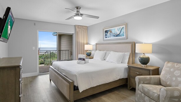 Hotelkamer met een bed, plafondventilator, schilderij, lamp, stoel en uitzicht op zee vanaf het balkon.