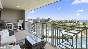 Een balkon met rieten meubels, een tafel met glazen blad en uitzicht op een resort met zwembad en strand.