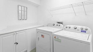Een wasserette met twee gestapelde wasmachines en drogers, een metalen droogrek, een raam met een rasterpatroon en een deur met een hangslot.