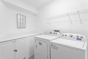 A laundry room with two stacked washers and dryers, a metal drying rack, a window with a grid pattern, and a door with a padlock.