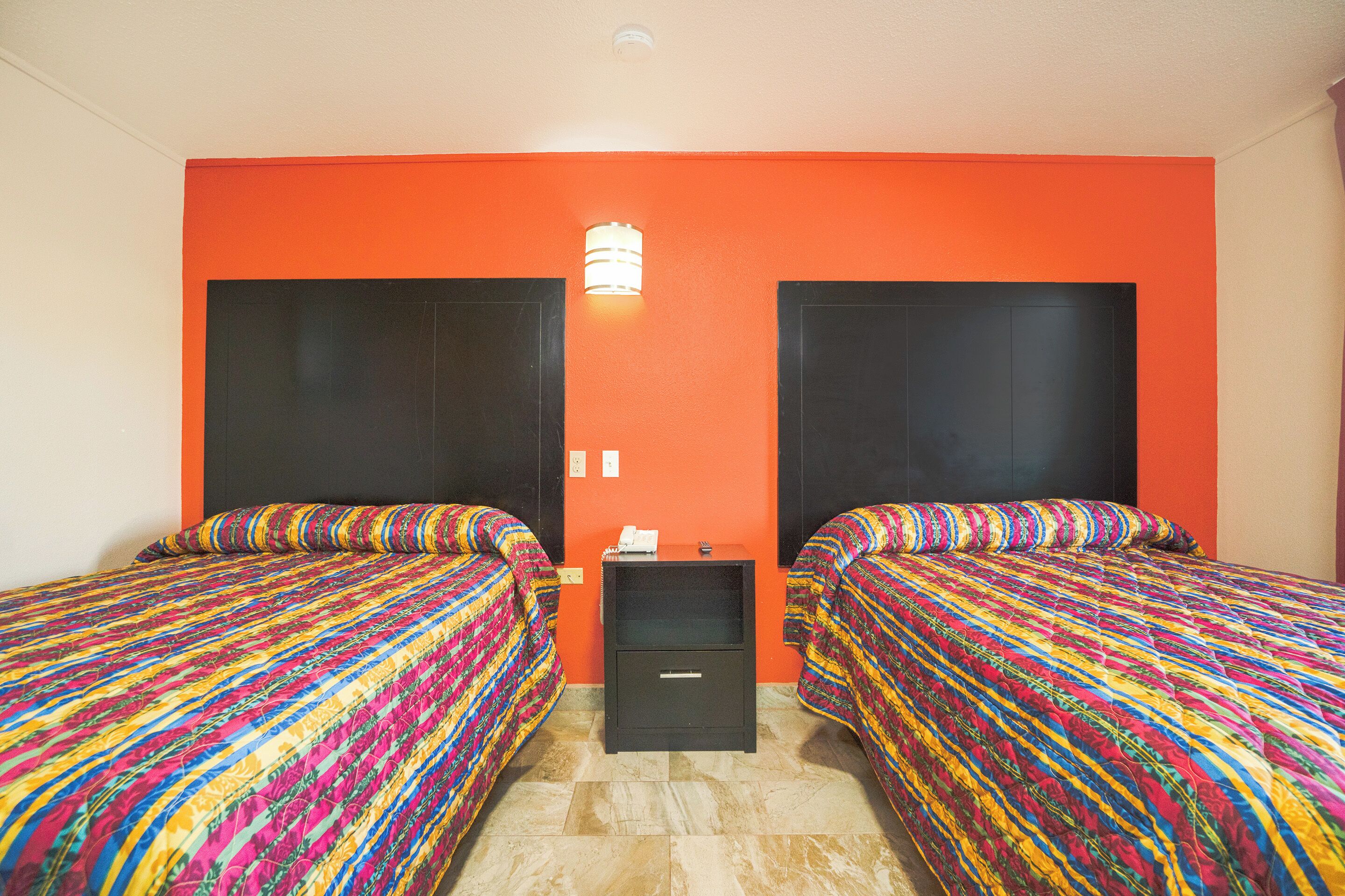 A room with two beds, each with colorful bedding, a small bedside table with a lamp, and a plain orange wall.