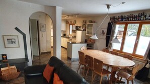 Uma sala de estar aconchegante com um sofá de couro preto, uma mesa de jantar de madeira com cadeiras e uma cozinha ao fundo.
