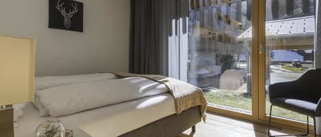 Een moderne hotelkamer met een groot bed, een bureau met lamp, een stoel en uitzicht op een gebouw en straat door grote ramen.