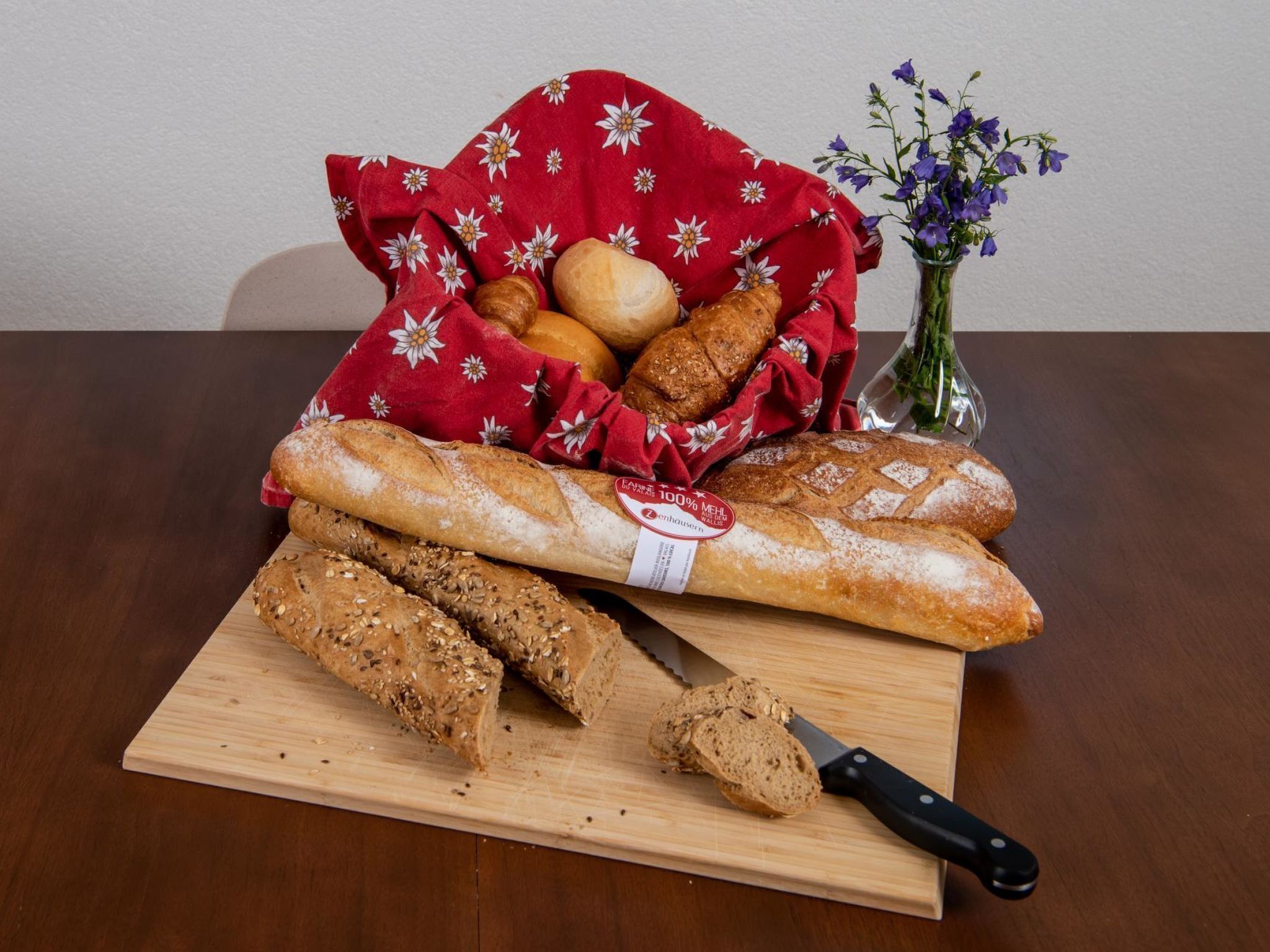 A wooden cutting board with sliced bread and a knife, a loaf of bread, and a basket of rolls on a table.