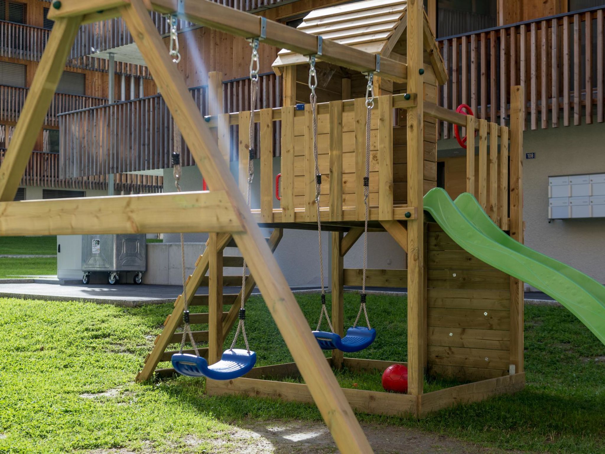 A wooden play structure with a slide and swings, set in a grassy area with a building in the background.