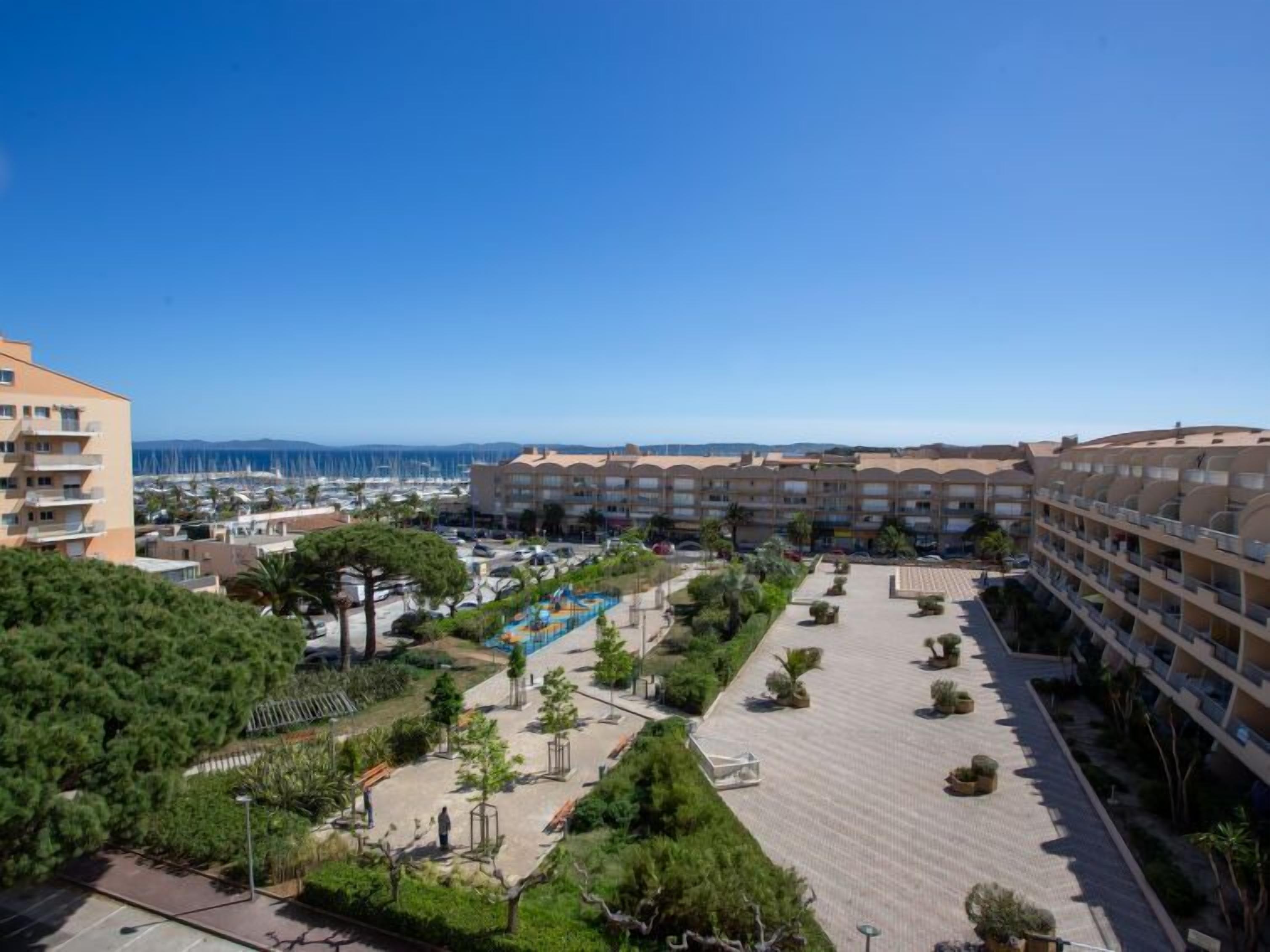 Un hôtel à étages, doté d’une cour intérieure avec des arbres et une piscine, et offrant une vue sur la mer et le port de plaisance en arrière-plan.