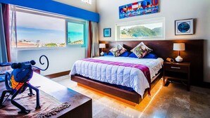 A bedroom with a large window, a bed with a blue and white bedspread, a nightstand with a lamp, and a painting on the wall.