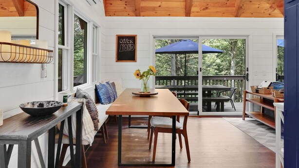 A dining area with a wooden table, chairs, and a bench. There is a window with a view of trees, a ceiling fan, and a sign that reads "OFFER ROOM."