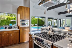 A modern kitchen with a built-in oven, a gas stove, and a large window overlooking a pool. - Aqua House - Luxury Midcentury Pool Retreat (Palm Springs)