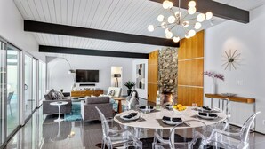 A modern living room with a dining area, large windows, a fireplace, and a chandelier.