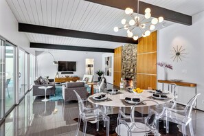A modern living room with a dining area, large windows, a fireplace, and a chandelier. - Aqua House - Luxury Midcentury Pool Retreat (Palm Springs)