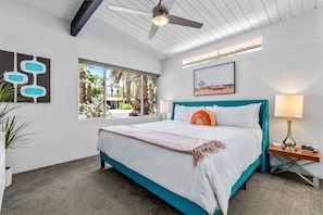 Quarto com cama, mesa de cabeceira, ventilador de teto, janela com vista para palmeiras e um quadro na parede.