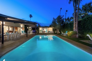 A swimming pool with lounge chairs, a covered patio area, and a well-lit building with visible windows and doors. - Aqua House - Luxury Midcentury Pool Retreat (Palm Springs)