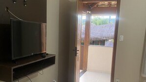 A room with a television on a wooden shelf, a white cabinet, and a door leading to an outdoor area with a thatched roof. - Pousada Cachoeira do Sol (Taquaraçu de Minas)