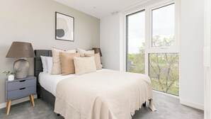 Quarto com uma janela grande, cama com várias almofadas, mesa de cabeceira com abajur e um quadro na parede.