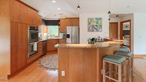 Una cocina amplia con armarios de madera, electrodomésticos de acero inoxidable, una isla central con taburetes y una obra de arte colorida en la pared.