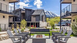 A modern outdoor seating area with Adirondack chairs and a fire pit, surrounded by a building with balconies and a mountain view in the background. - River West 924 - Silverthorne Condo with River Views. (Silverthorne)