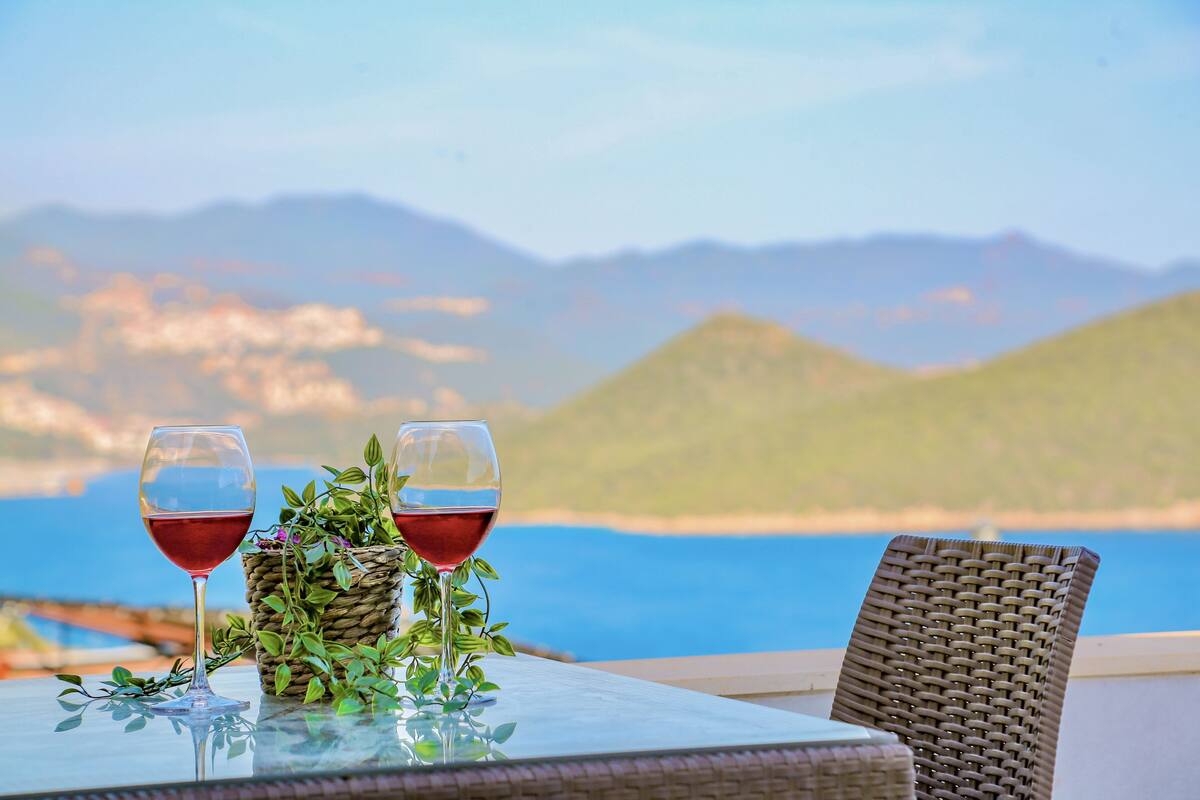 Deux verres à vin sont posés sur une table, à côté d’un panier en osier et de quelques plantes vertes. De là, on peut admirer un paysage magnifique, avec des montagnes et un plan d’eau en toile de fond.
