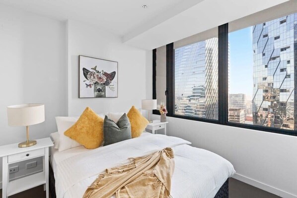 A modern hotel room with a large bed, white walls, and a city view through a large window.