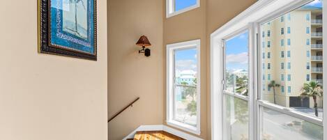 A corner room with a wooden floor, a framed picture on the wall, a lamp, and large windows with a view of a building and palm trees.