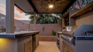 Un área de cocina al aire libre con una parrilla empotrada, un ventilador de techo y vistas a un muro de ladrillo y palmeras.