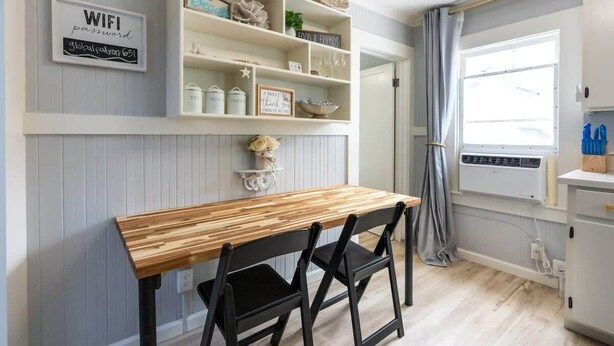 A dining area with a wooden table and black chairs, a white shelf with decorative items, and a window with curtains. - Capitola Village Bungalow - 2 Blocks from the Sand (Capitola)