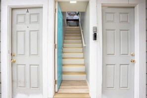 A narrow staircase with a teal door on the left and a white door on the right, leading to a landing area with a small table and chair.