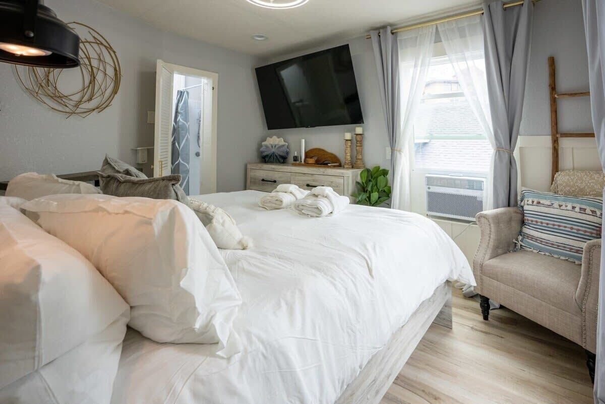 A bedroom with a large bed, a TV mounted on the wall, a bedside table with a lamp, and a window with curtains.