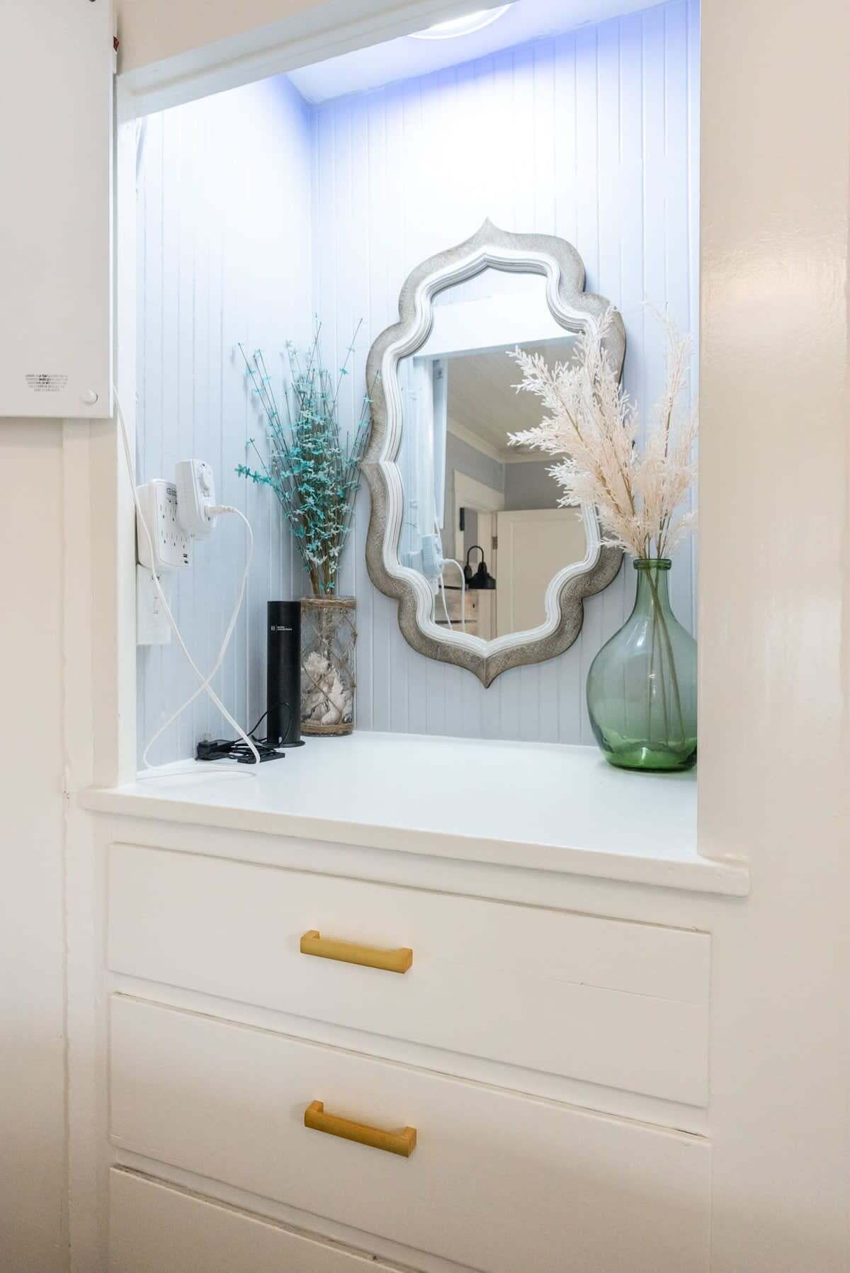 Une coiffeuse blanche avec un miroir, deux tiroirs et un vase contenant des fleurs séchées.